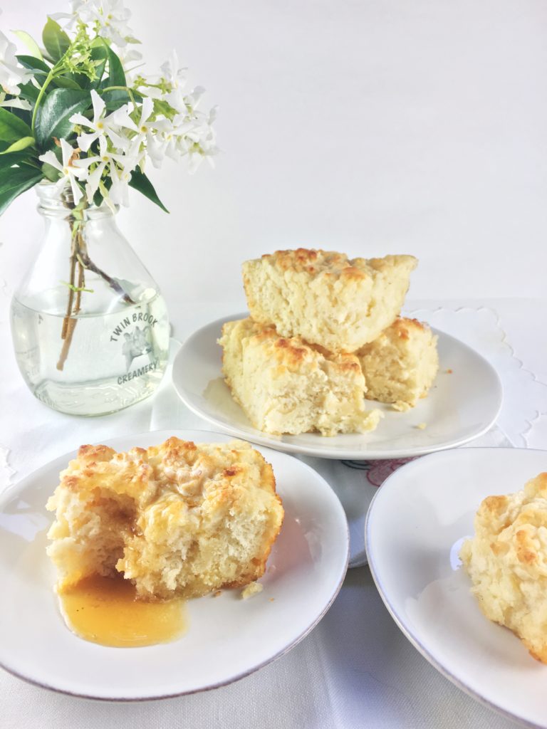 Shows buttermilk biscuit covered with honey, with more biscuits on plates around it and a vase of white flowers