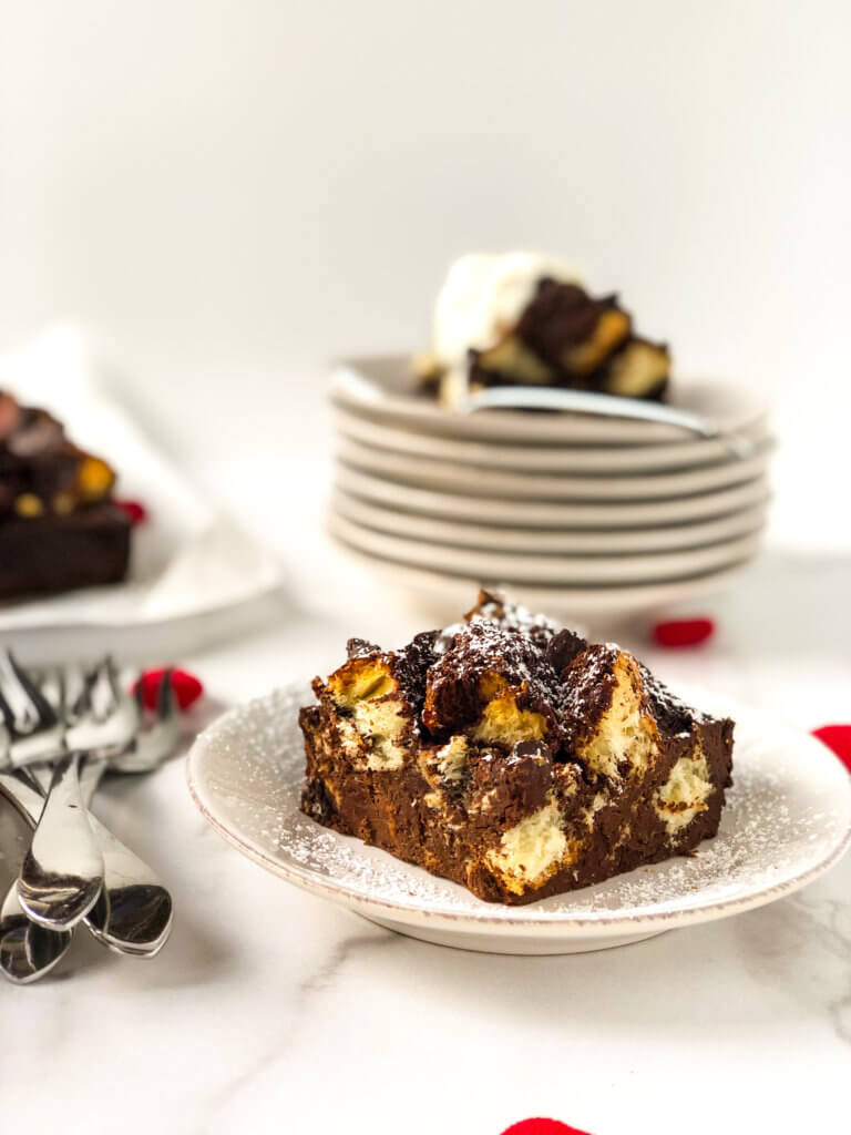 Shows slices of bread pudding, one with whipped cream one with powdered sugar, and a glimpse of the pan full of bread pudding