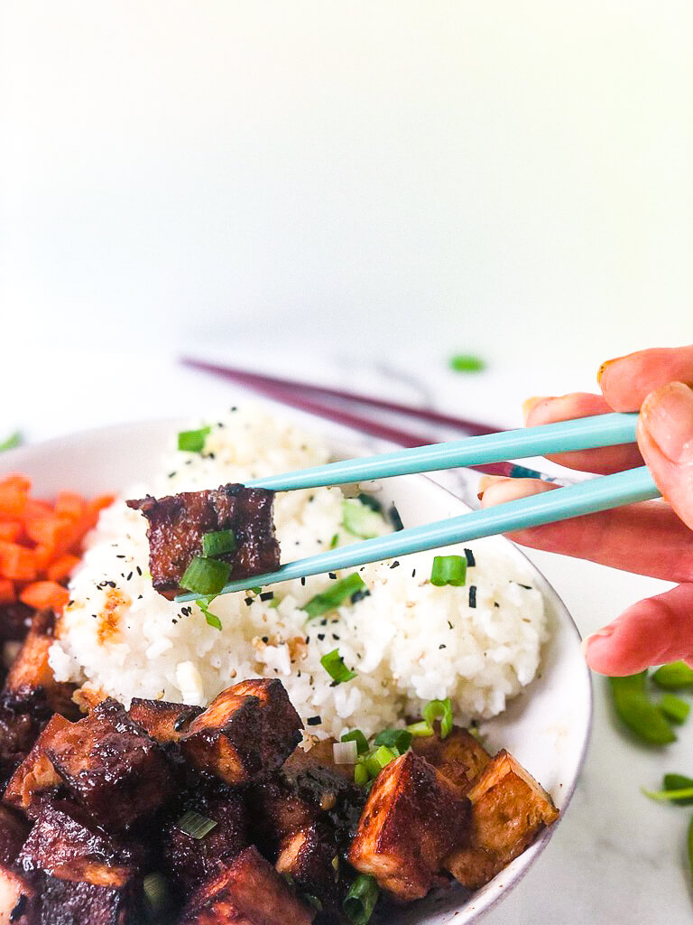 Shows cube of marinated crispy tofu in chopsticks hovering over rice bowl