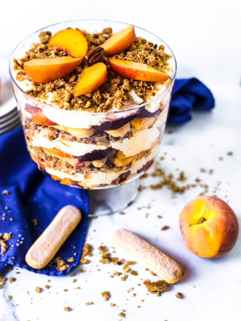 Featured Photo for Peach Crisp Trifle showing the trifle from above with scattered ingredients including lady fingers, baked crisp and fresh peaches. blue napkin drapes in white background