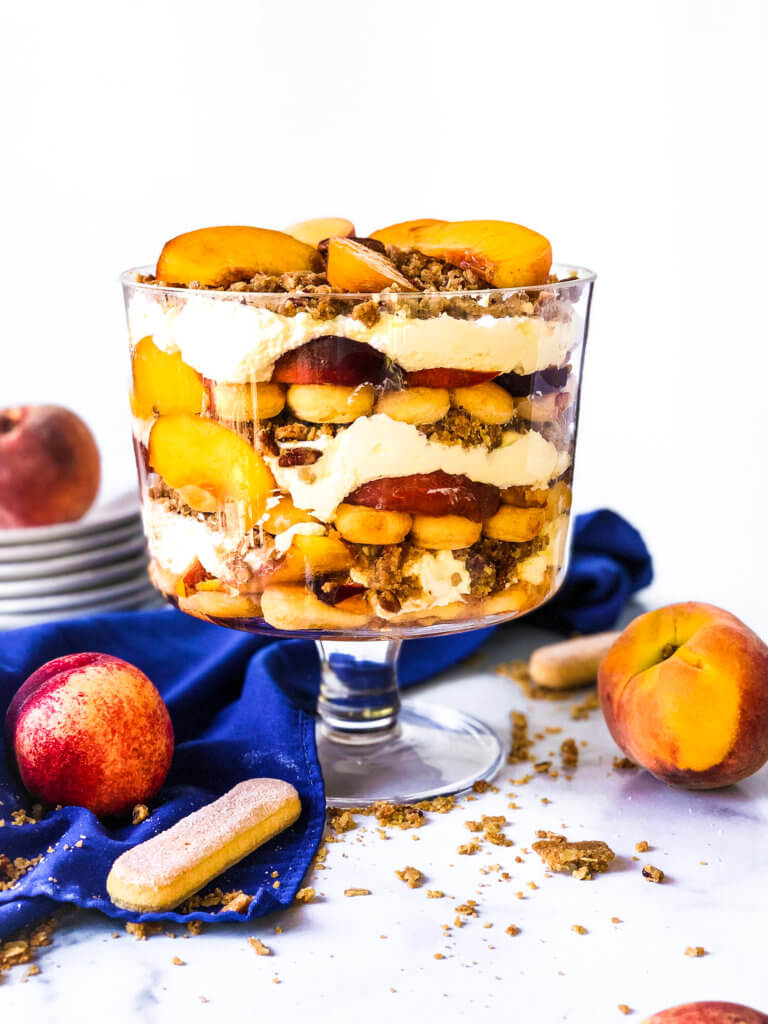 Easy Peach Crisp Trifle photo showing the trifle from above with scattered ingredients including lady fingers, baked crisp and fresh peaches. blue napkin drapes in white background