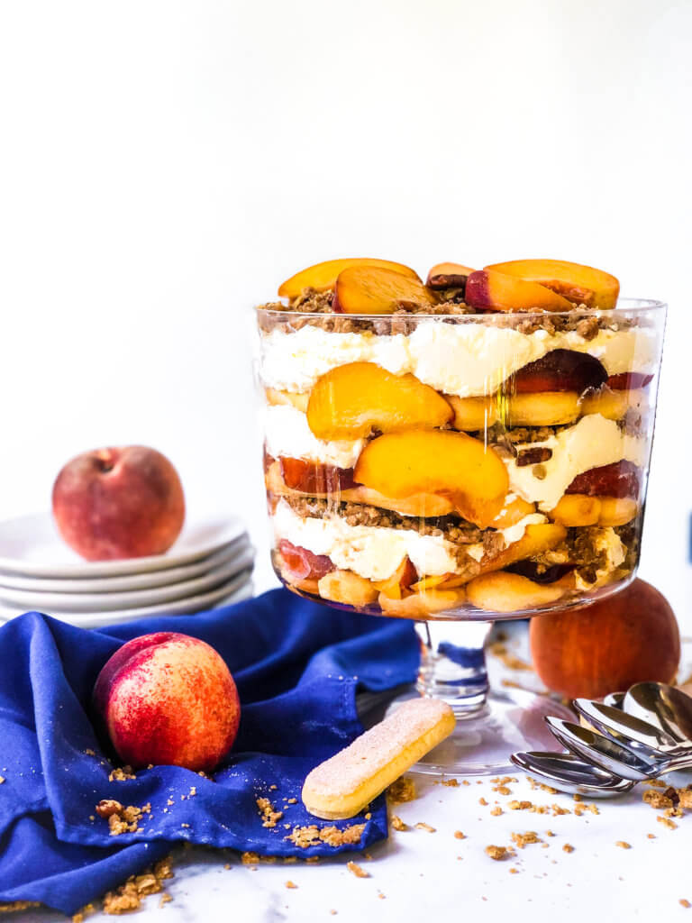 12-layered Peach Crisp Trifle showing the trifle from above with scattered ingredients including lady fingers, baked crisp and fresh peaches. blue napkin drapes in white background