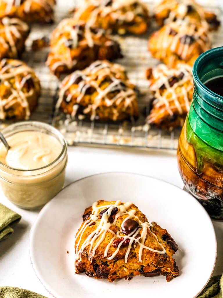 Closeup of Pumpkin Chocolate Chip Scone on white plate with a small jar of Brown sugar sauce and tray of more drizzled scones in the background