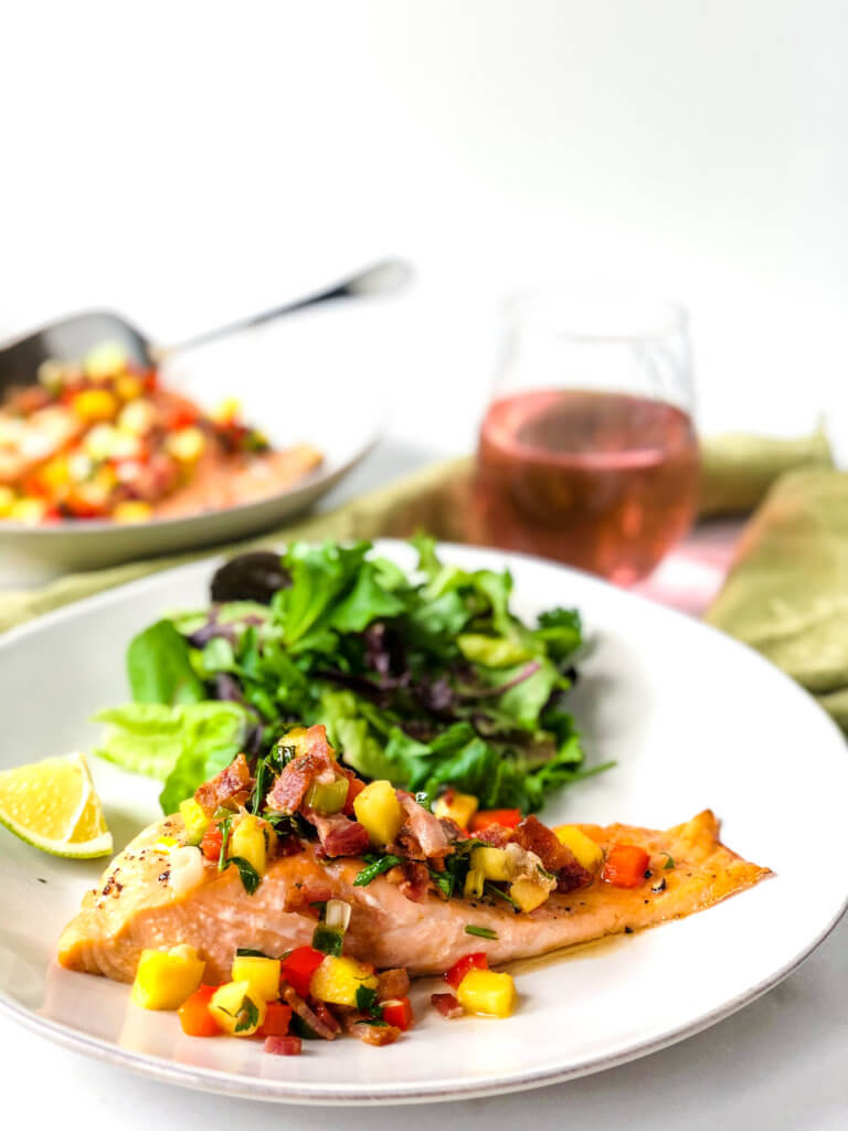 Fillet of salmon topped with pineapple bacon salsa on plate with green salad. In the background a glass of rose and bowl with more salsa-covered salmon