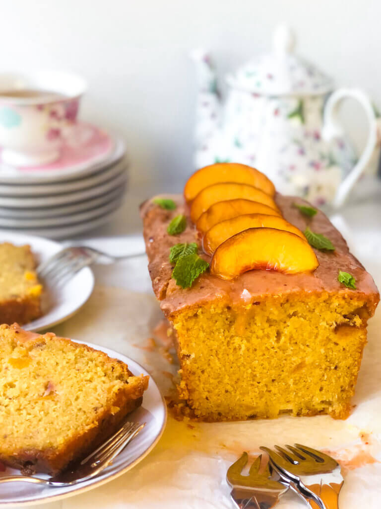 Shows peach poundcake loaf with a line of peach slices on top with a few mint leaves. Several slices on plates next to the cake. A teacup and teapot in the background
