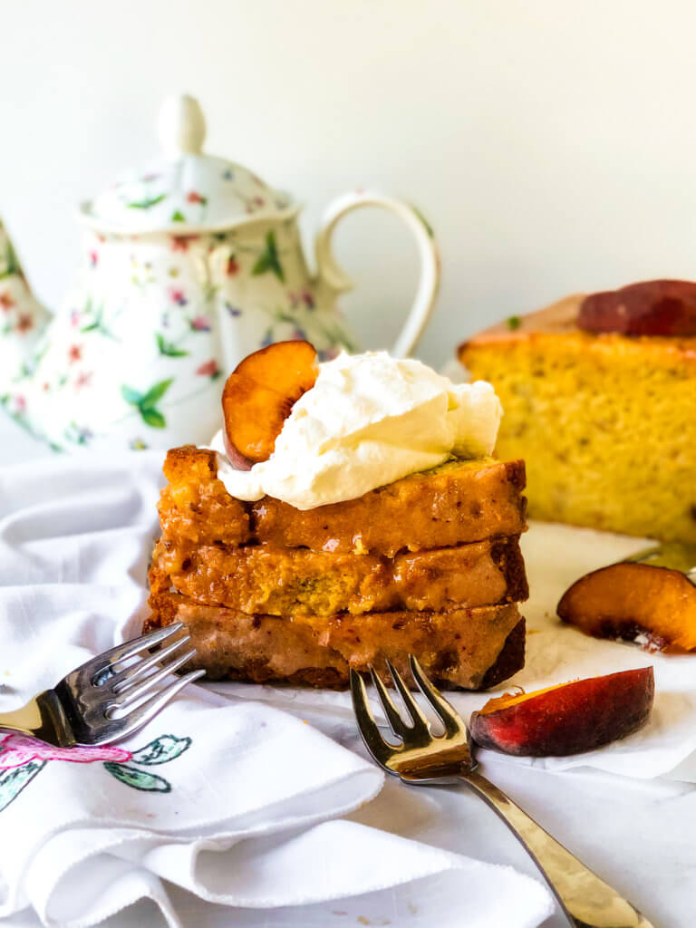 Featured image. Shows 3 stacked slices of peach lime pound cake, topped with whipped cream and peach slices. In the background is a teapot and the remainder of the loaf cake. White flowered napkin and forks strewn about white background