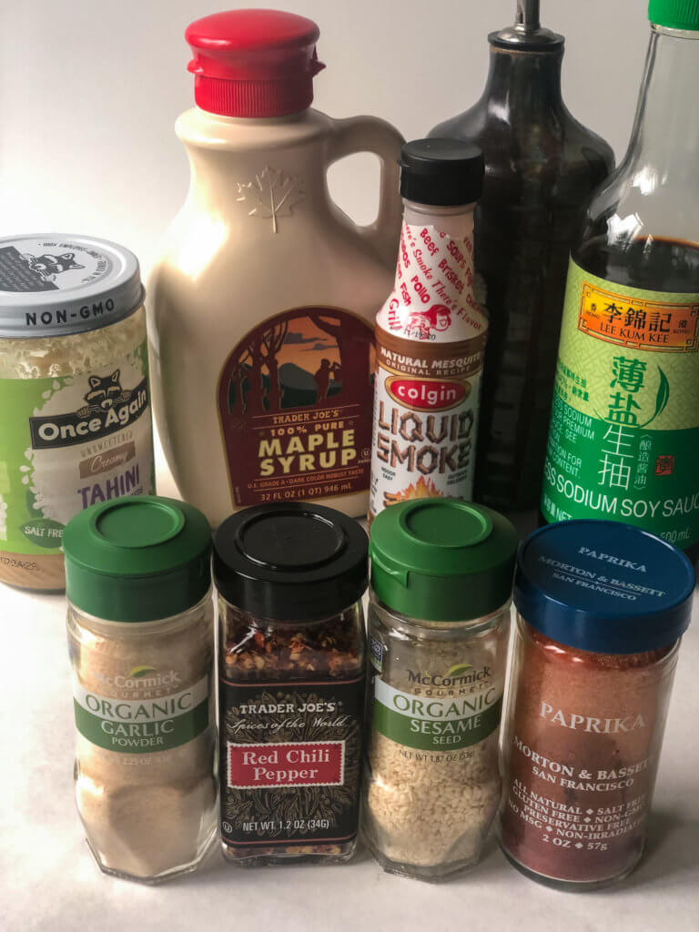 Process shot showing ingredients on white background: (back row) Tahini, Maple Syrup, liquid smoke, olive oil, low-sodium soy sauce. (front row) Garlic powder, Crushed Red Pepper, sesame seeds, paprika