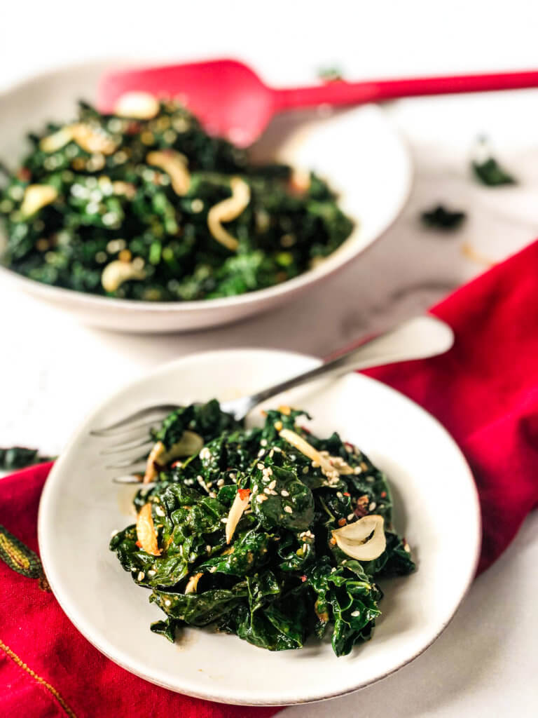 Closeup of small plate of Smoky Kale, with serving dish in the background. A red spoon in the serving dish. the small plate setting over a red napkin.