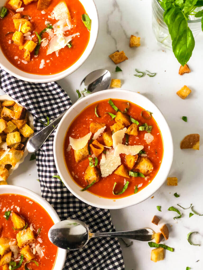 overhead view Shows 3 bowls of tomato white bean soup with croutons, Parmesan shavings and shredded basil, with bowl of croutons to the side of the middle bowl