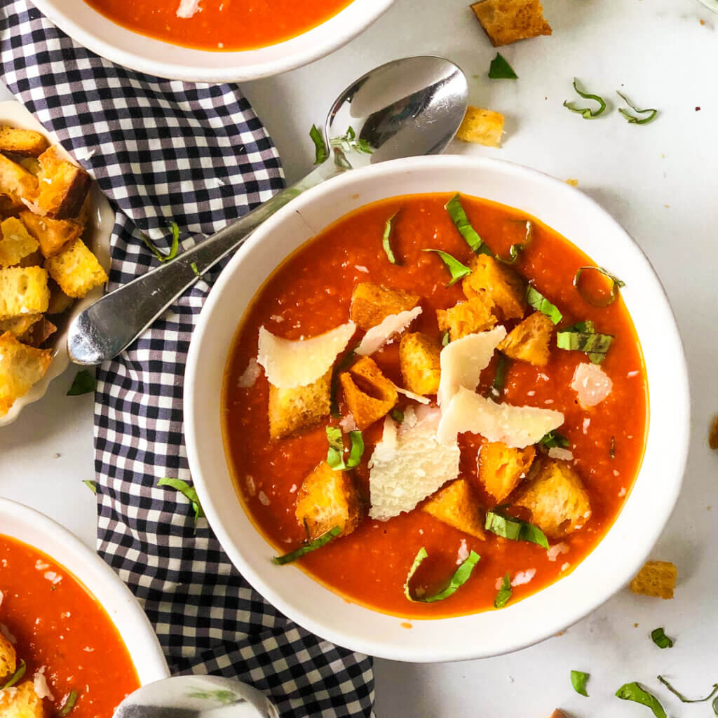 Overhead closeup of bowl of tomato white bean soup, topped with golden croutons, shreds of parmesan and bits of basil, spoon rising on black and white gingham napkin