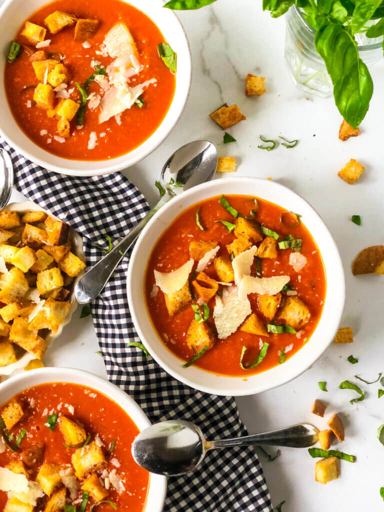 overhead view Shows 3 bowls of tomato white bean soup with croutons, Parmesan shavings and shredded basil, with bowl of croutons to the side of the middle bowl