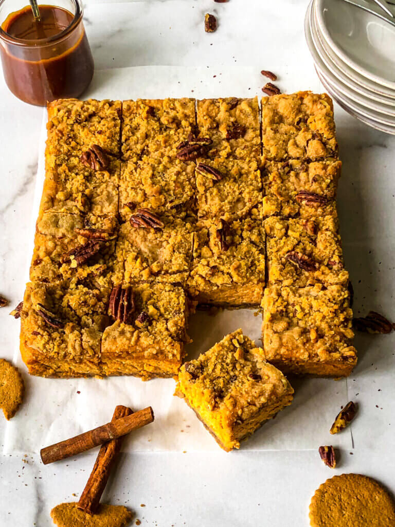 Shows Sweet Potato Cheesecake bars against white background, with scattered gingerbread cookies, cinnamon and chopped pecans