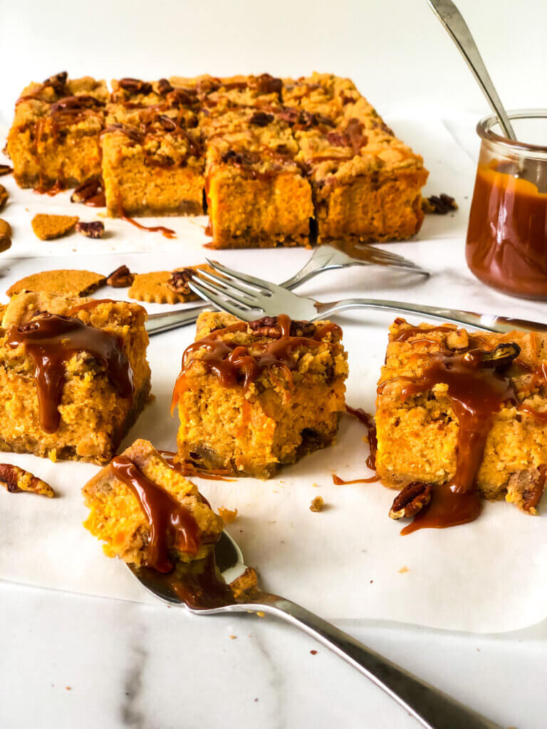 Blog photo Shows 3 slices of Sweet Potato Cheesecake in foreground, a bite taken out of one with a fork, remaining bars and jar of caramel in background