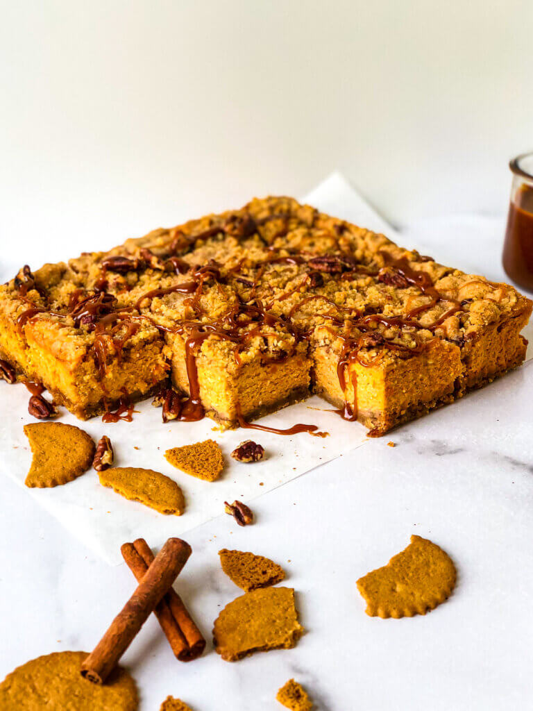 Blog photo Shows Sweet Potato Cheesecake bars against white background, with scattered gingerbread cookies, cinnamon and chopped pecans