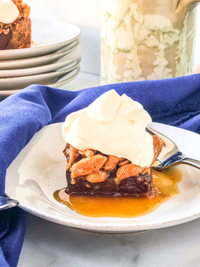 Closeup of Chocolate Peanut pie bar layers, including chocolate ganache over a cookie crust, toped with peanuts and whipped cream, sitting in caramel sauce, Mug and blue napkin in background