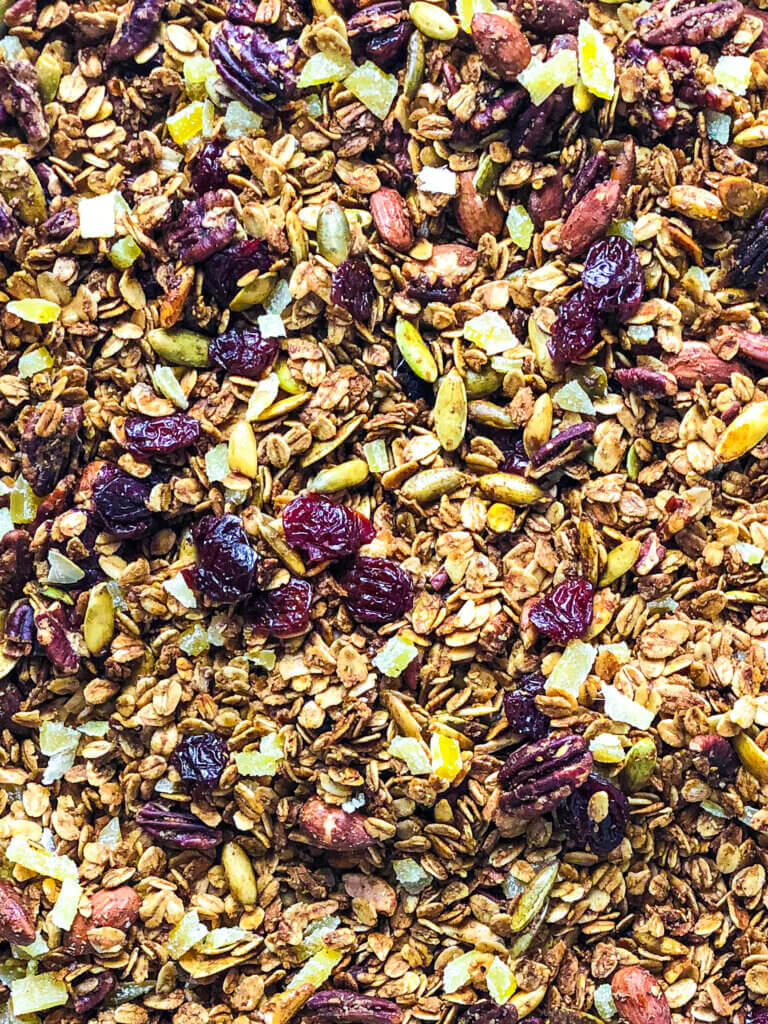 Process Shot Homemade Gingerbread granola shows overhead close-up baked golden granola with dried cranberries and bits of candied ginger