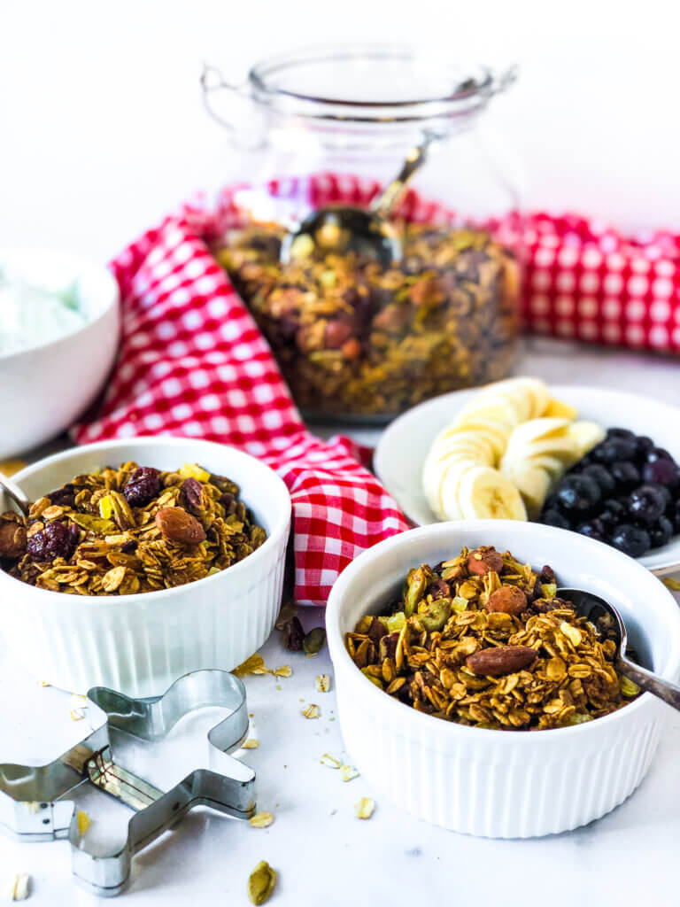 Shows white background with a jar half-full of granola and a red gingham napkin. In the foreground there are two small bowls, full of granola, a plate with berries and bananas and a gingerbread cookie cutter