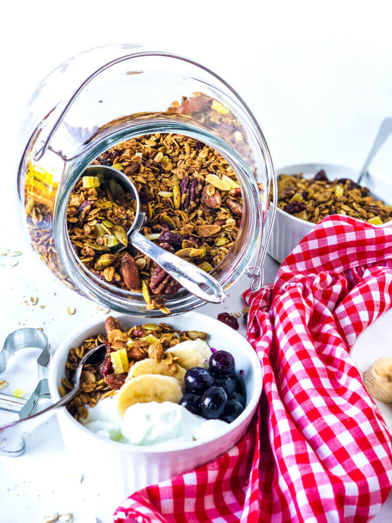 Blog image showing jar of gingerbread granola tipped over into a white bowl with granola, yogurt, blueberries and banana slices. Red gingham napkin at right side and gingerbread man cookie cutter at left