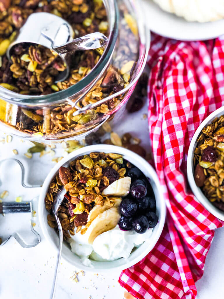 Overhead shot of glass jar holding granola, two bowls of granola, yogurt and fruit, a red gingham napkin and gingerbread man cookie cutter, all around a red gingham napkin