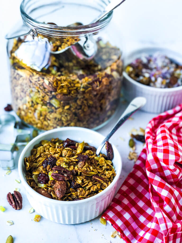 Blog photo showing jar of granola, and ramekin in front and at the side, each containing granola. Toward front is red gingham napkin