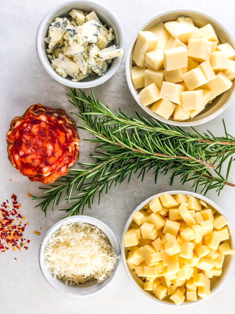 Process shot making white pizza shows Marbled white background with bowls of gorgonzola cheese, mozzarella, provolone, parmeggiano, red chili flakes, salmi, and rosemary