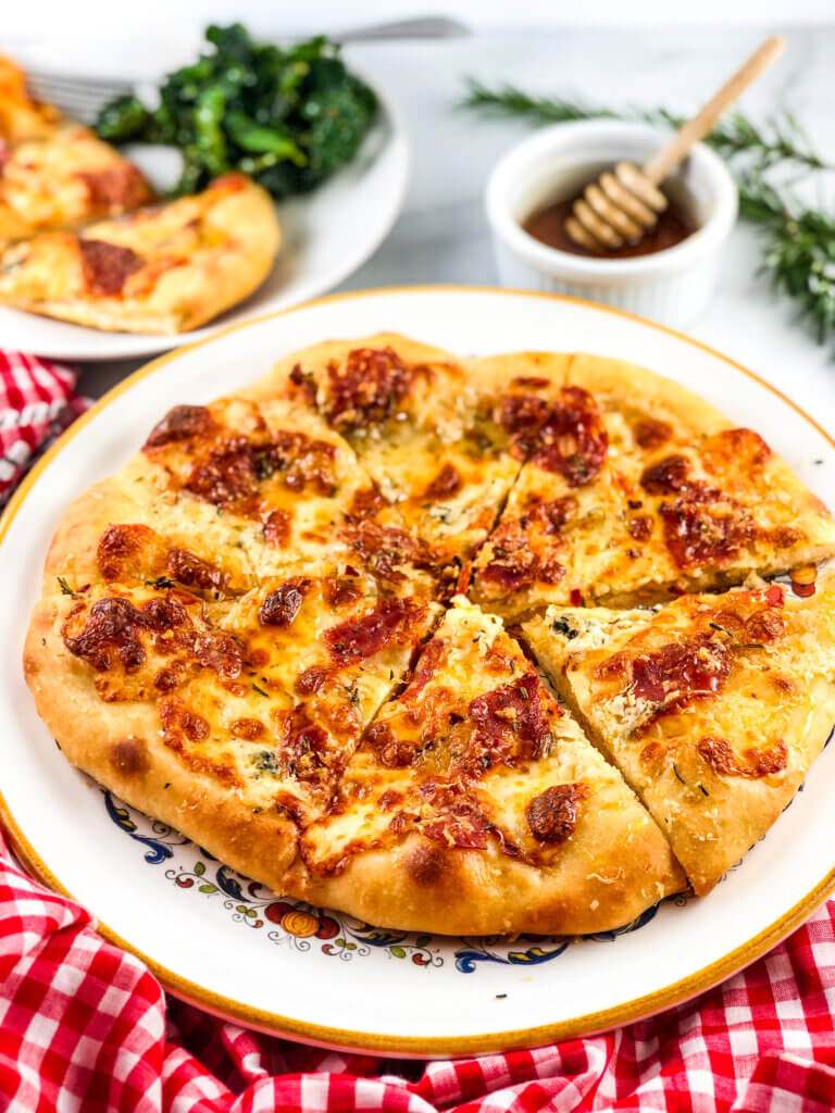 Shows fully baked, golden brown-crusted, melty cheese, salami all over pizza. Behind the pizza are a pot of honey for drizzling and a plate with pizza and a green salad. Red/white gingham napkin in the foreground