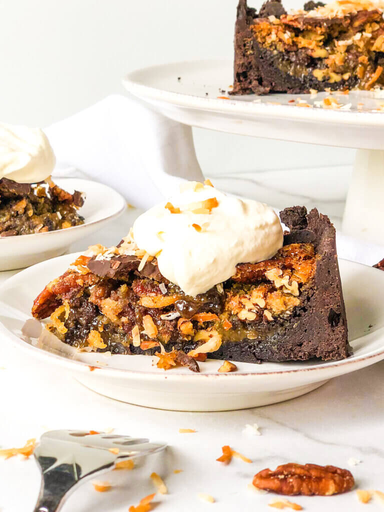 Blog post photo showing Close-up, head-on view of slice of chocolate pecan pie tart with dollop of whipped cream. In background is cake stand with remaining pie. All on white background with fork, scattered toasted coconut and pecans.