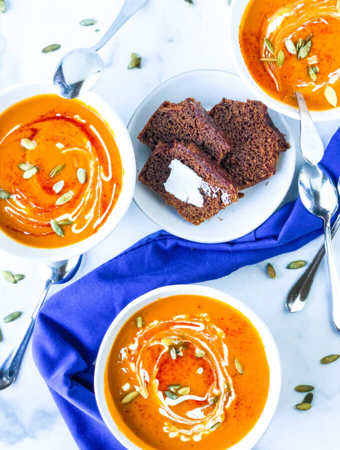overhead view of 3 scattered bowls of smoky sweet potato butternut soup with a plate of buttered brown bread in the middle. scattered spoons and pepitas with a blue napkin. All on marble slab