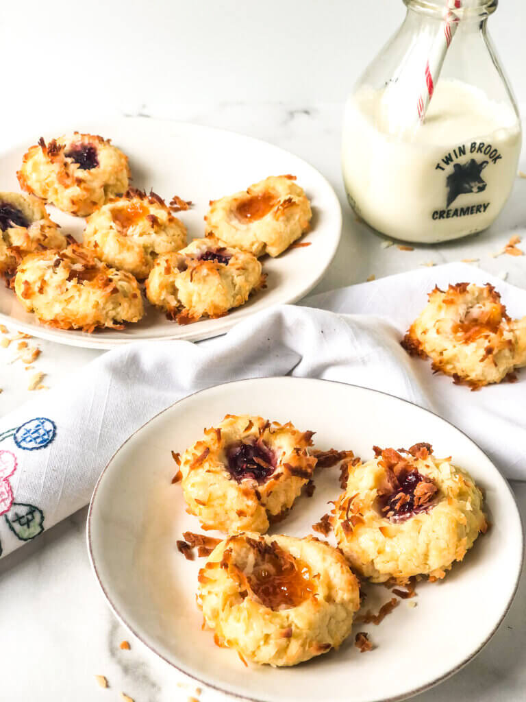 Blog post photo showing an plate of apricot and raspberry coconut-coated thumbprint cookies, with one missing. Bottle of milk with straw, plates and cookie on white napkin in the white background