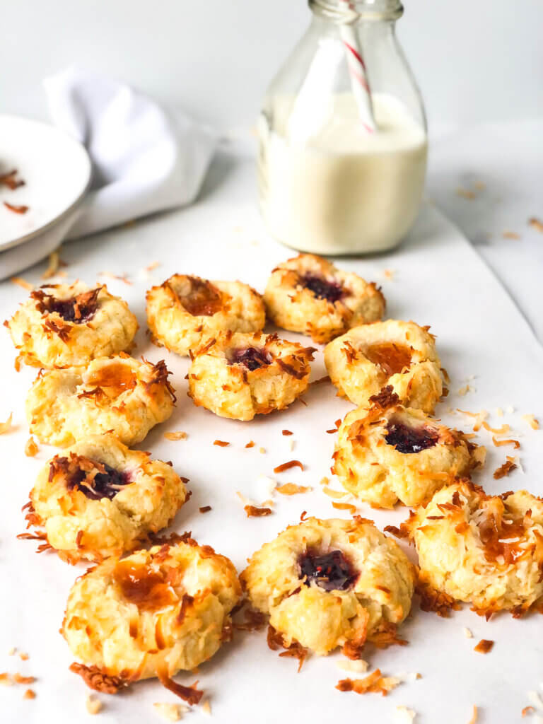 Blog post photo showing an array of apricot and raspberry coconut-coated thumbprint cookies, with one missing. Bottle of milk with straw, plates and cookie on white napkin in the white background