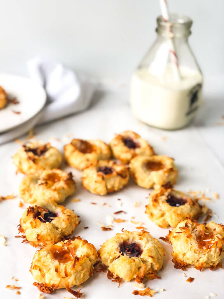 Blog post photo showing an array of apricot and raspberry coconut-coated thumbprint cookies, with one missing. Bottle of milk with straw, plates and cookie on white napkin in the white background