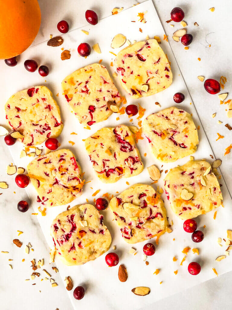 Blog photo showing how to make cranberry orange almond shortbread cookies 3 rows and 3 columns of square cranberry orange almond shortbread cookies with scattered cranberries and an orange showing 