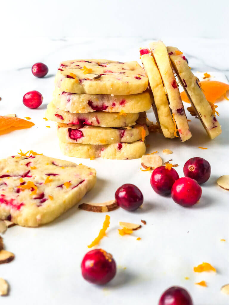 blog photo showing Stack of 4 square Almond Shortbread cookies with orange and cranberries, with 3 more stacked sideways against it and another lying in front with scattered orange zest and cranberries