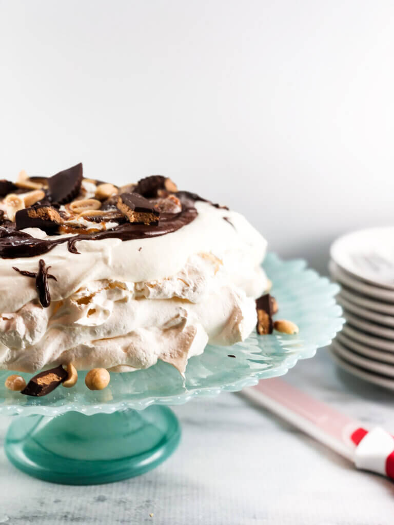 Pavlova covered in mascarpone whipped cream and topped with chocolate sauce, peanut butter cups and peanuts.
