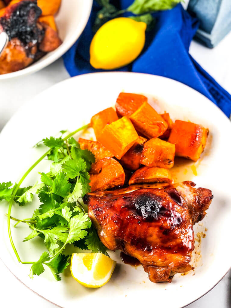 Blog photo showing overhead view of plate of roasted sheet pan maple chicken and sweet potatoes with a lemon wedge, a dish of maple sauce, a lemon and a bowl containing more chicken and sweet potatoes