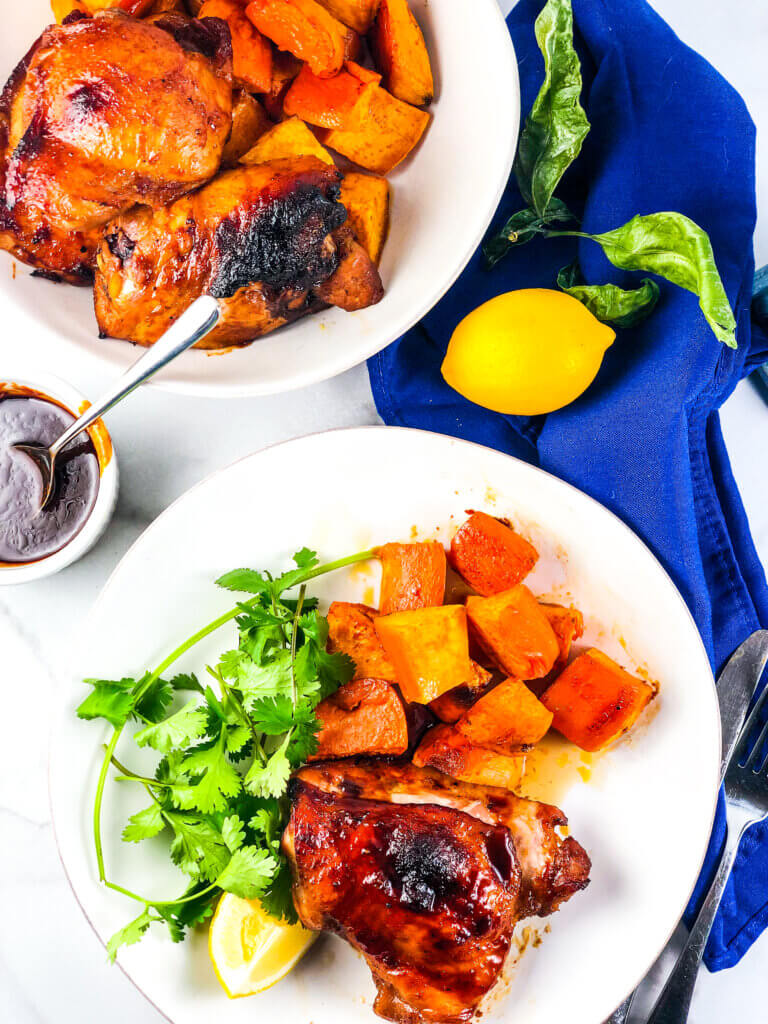 Blog photo showing overhead view of plate of roasted sheet pan maple chicken and sweet potatoes with a lemon wedge, a dish of maple sauce, a lemon and a bowl containing more chicken and sweet potatoes