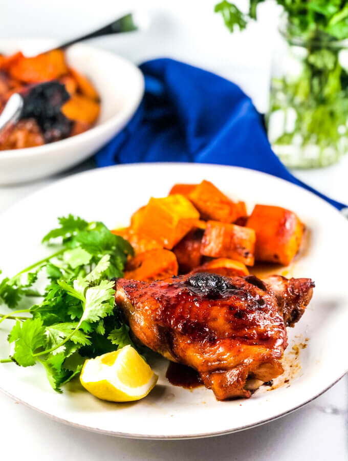 Blog photo showing plate of roasted sheet pan maple chicken and sweet potatoes with a lemon wedge. Blue napkin and vase of greens in background