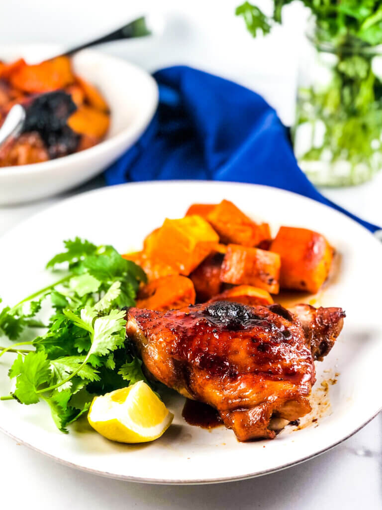 Blog photo showing plate of roasted sheet pan maple chicken and sweet potatoes with a lemon wedge. Blue napkin and vase of greens in background