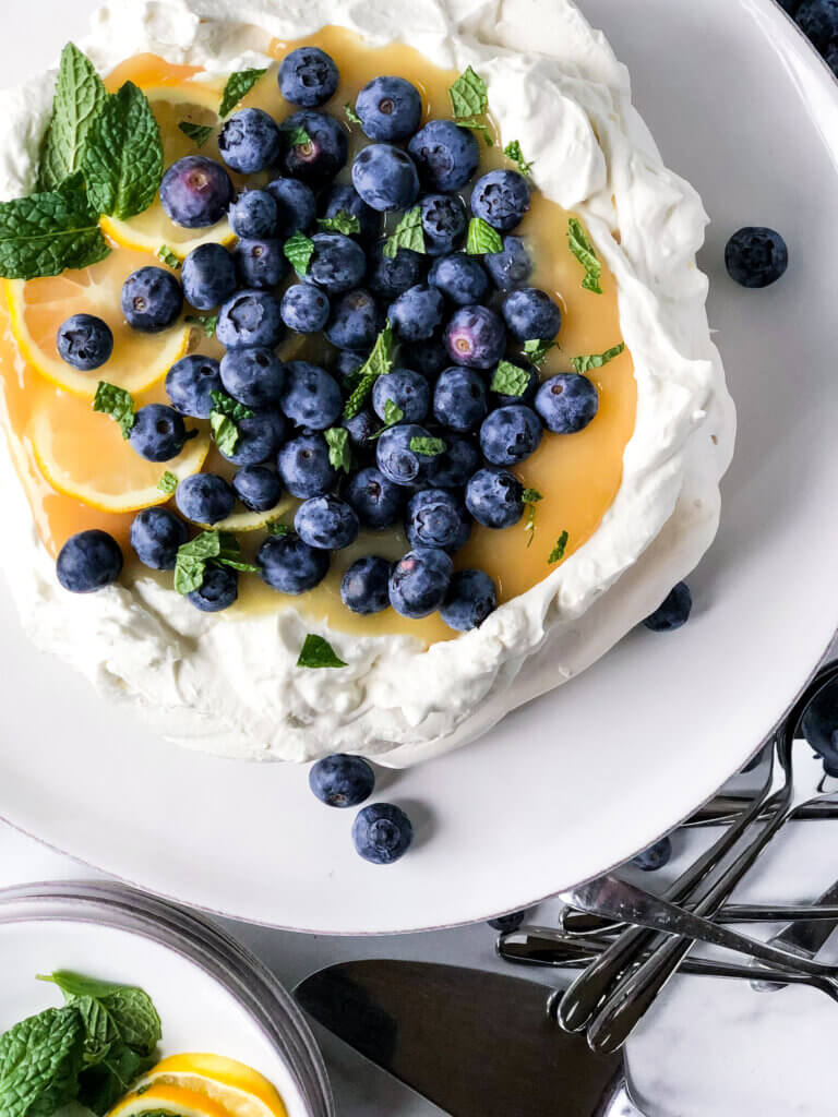 Pavlova with mascarpone cream and filled with lemon curd, sprinkled with blueberries on a white cake plate. Bowl of blueberries at bottom left.