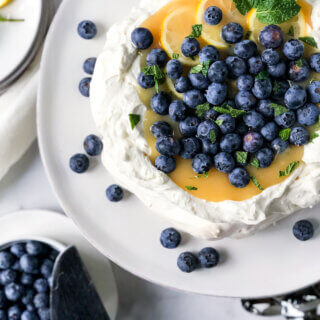 Pavlova with mascarpone cream and filled with lemon curd, sprinkled with blueberries on a white cake plate. Bowl of blueberries at bottom left.