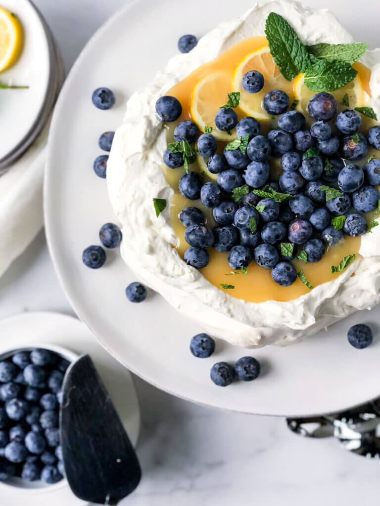 Pavlova with mascarpone cream and filled with lemon curd, sprinkled with blueberries on a white cake plate. Bowl of blueberries at bottom left.