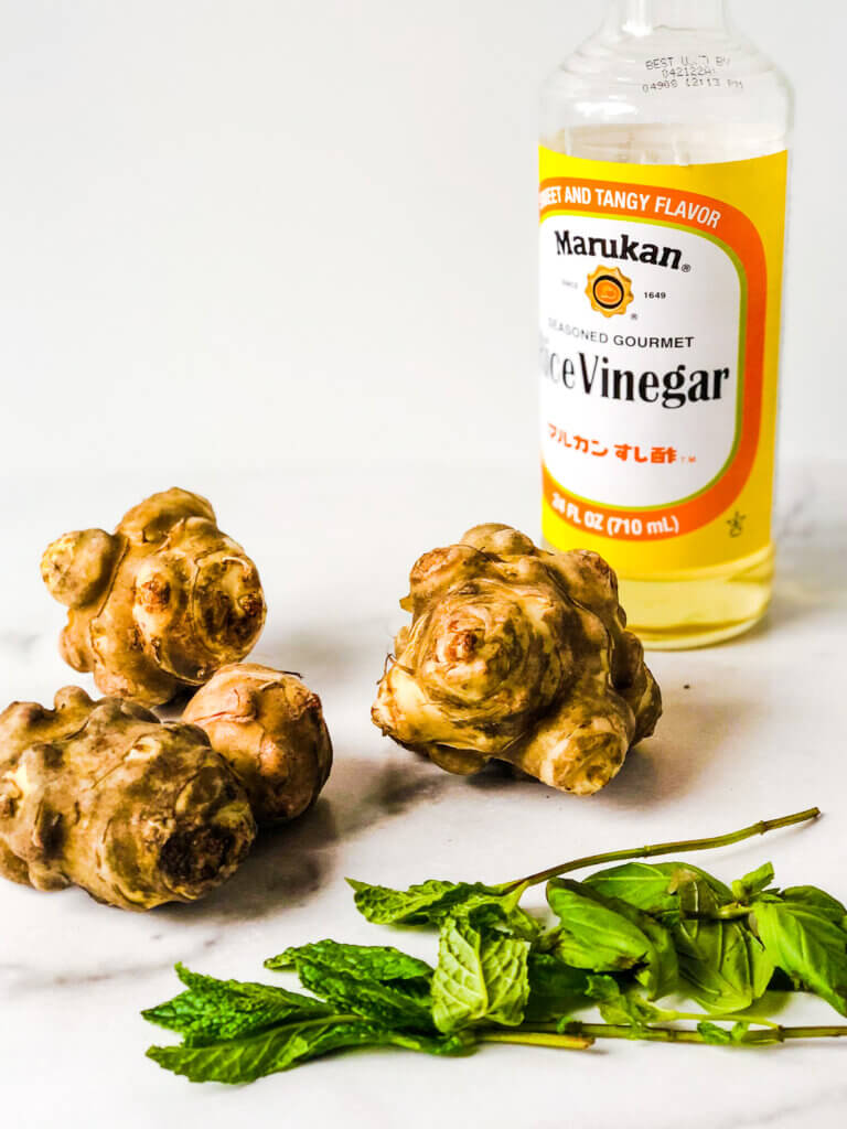 Process shot showing 4 sunchokes, bottle of seasoned rice vinegar, basil and mint leaves