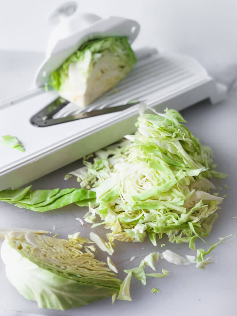 Shows mandolin with section of cabbage and a pile of shaved cabbage in the front