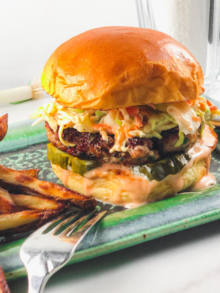 Closeup view of Ginger Pork Burger, with sauce dripping down the burger, slaw and pickles topping the burger, all in a bun. 