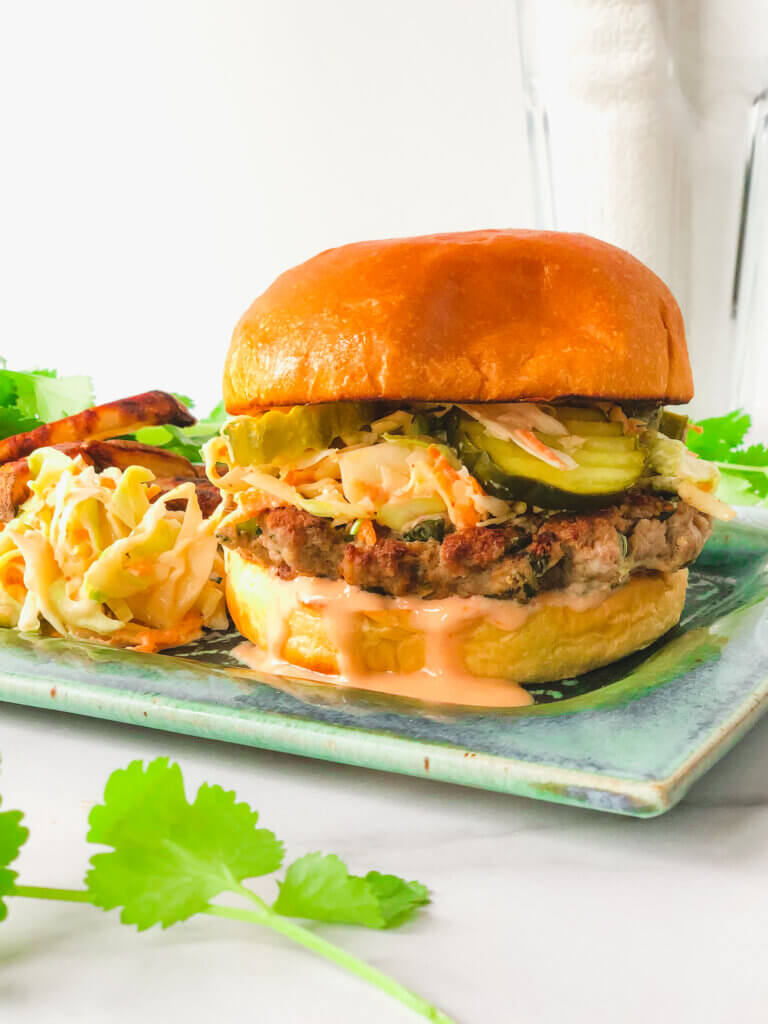 Featured Blog photo shows closeup of ginger pork burger topped with slaw, pickles and sweet and spicy dressing on bun. Sits on plate with extra slaw and crispy fries